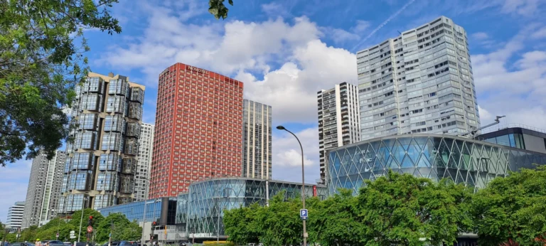 Residential towers and a glass-clad shopping centre in an urban district, under a cloudy blue sky
